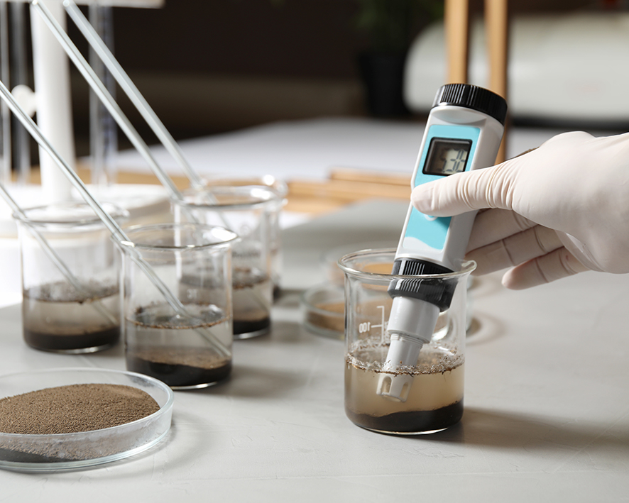 Meranie pH pomocou pH metra hnedej sypkej zmesy v sklenej nádobke na bilom stole v prostredí chemického štúdia.